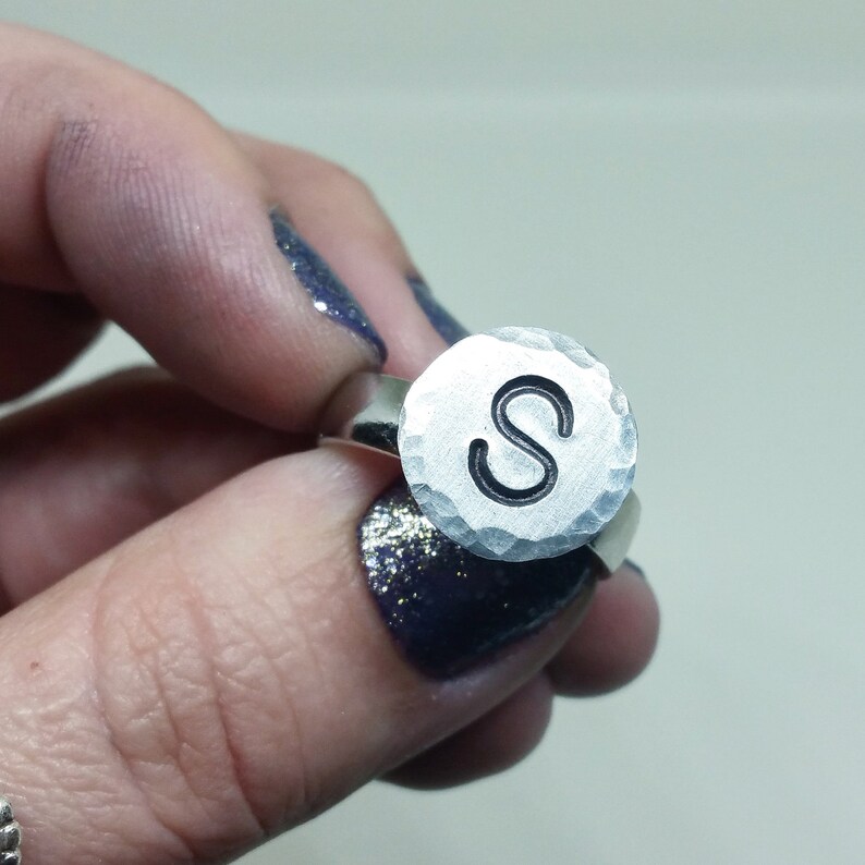 Op de afbeelding: Een zilveren ring met een gehamerde, cirkelvormige voorkant. De voorkant heeft de letter "S" in een vetgedrukte, zwarte letter. De ring wordt vastgehouden door een hand met donkerblauwe nagellak.