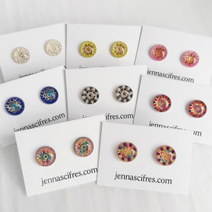 Puede incluir: Una colección de pendientes de botón de varios colores y diseños, expuestos en tarjetas blancas. Los pendientes presentan diseños florales y geométricos en tonos azules, rosas, amarillos y más. La dirección del sitio web jennascifres.com está impresa en las tarjetas.