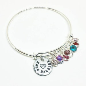 May include: Silver bangle bracelet with a charm that says "Nana Barbara" with a heart symbol. The bracelet has multiple dangling charms with colorful gemstones.