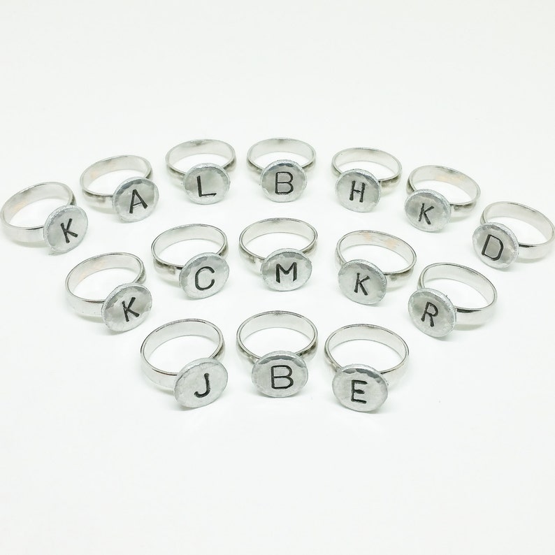 Op de afbeelding: Een verzameling zilverkleurige ringen, elk met een gehamerde, cirkelvormige face met een enkele zwarte letter. De letters zijn A, B, C, D, E, H, J, K, L, M en R. De ringen zijn gerangschikt op een witte achtergrond.
