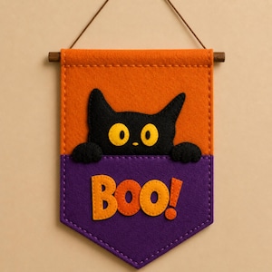 May include: A Halloween-themed felt banner with a black cat peeking out from behind a purple pocket. The banner has an orange background and the word "Boo!" in orange letters. A wooden dowel and twine are at the top for hanging.