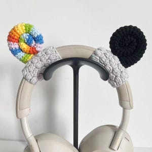 May include: A pair of white headphones with custom crochet ear covers. One cover is a spiral of rainbow colors, the other is black. The headband has gray crochet accents. The headphones are on a black stand.