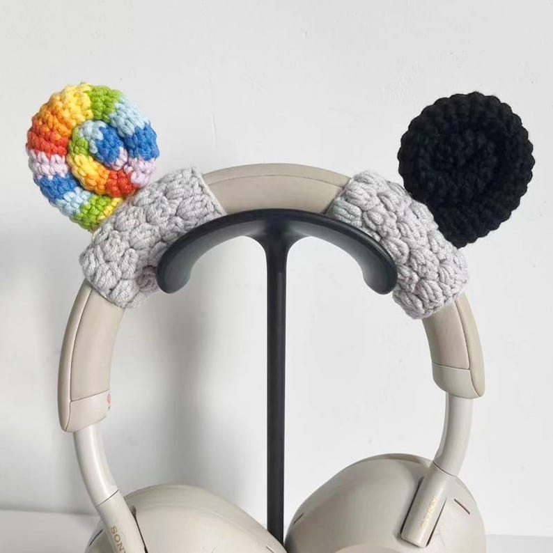 May include: A pair of white headphones with custom crochet ear covers. One cover is a spiral of rainbow colors, the other is black. The headband has gray crochet accents. The headphones are on a black stand.