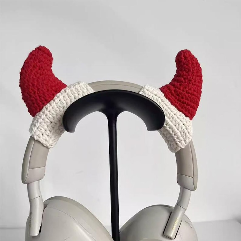 May include: A pair of headphones with red and white crocheted devil horn covers. The horns are red with white trim, and the headphones are light gray. The headphones are on a black stand against a white background.