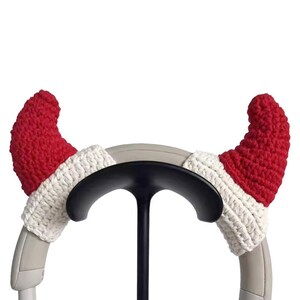 May include: A pair of red and white crocheted devil horn covers for headphones. The horns are red with a white trim, designed to fit over the earcups. The headphones are supported by a black stand.