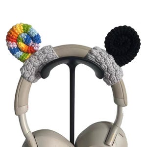 May include: Headphones with gray ear covers and decorative crochet accents. One accent is a rainbow swirl, the other is a black circle. The headphones are on a black stand, showcasing the colorful and unique design.