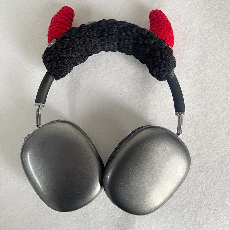 K&ouml;nnte beinhalten: Graue Kopfh&ouml;rer mit einem schwarzen geh&auml;kelten Stirnband mit zwei roten geh&auml;kelten H&ouml;rnern. Die Kopfh&ouml;rer haben silberne Akzente und ein modernes Design, ideal f&uuml;r Musikliebhaber.