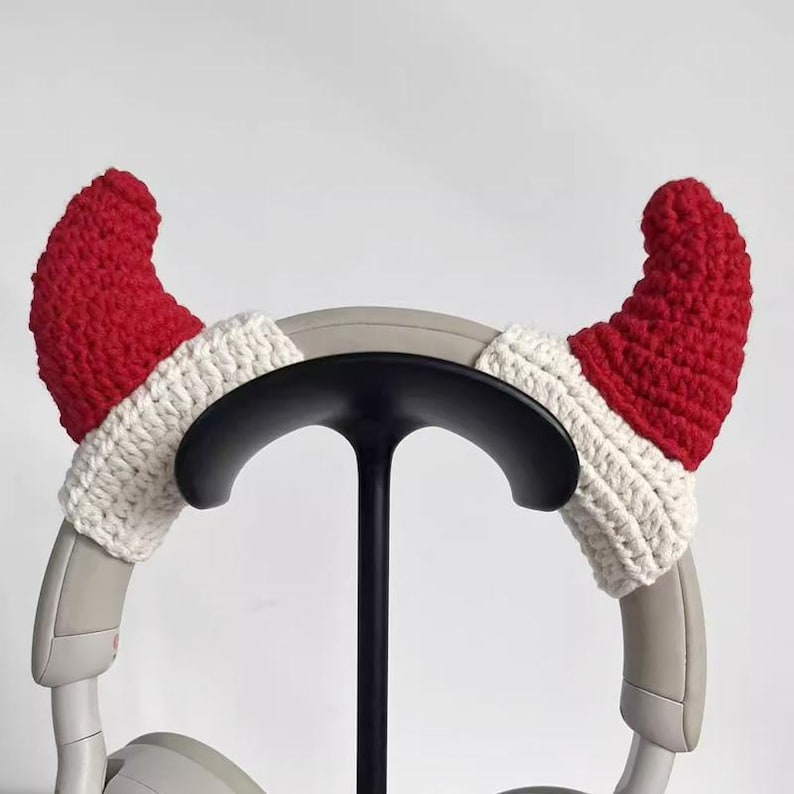 May include: A pair of red and white crocheted Santa hat ear covers for headphones. The ear covers are designed to fit over the earcups of the headphones, adding a festive touch. The headphones are gray and are on a black stand.