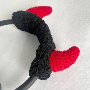 K&ouml;nnte beinhalten: Ein schwarzes geh&auml;keltes Stirnband mit zwei roten geh&auml;kelten H&ouml;rnern. Die herzf&ouml;rmigen H&ouml;rner sind an dem schwarzen Stirnband befestigt. Ein lustiges Accessoire f&uuml;r Kost&uuml;me oder Themenveranstaltungen.