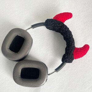 K&ouml;nnte beinhalten: Graue Kopfh&ouml;rer mit einem geh&auml;kelten Teufelshorn-Stirnband in Schwarz und Rot. Das Stirnband ist schwarz mit zwei roten H&ouml;rnern. Die Kopfh&ouml;rer haben graue Ohrmuscheln und ein silbernes Band.