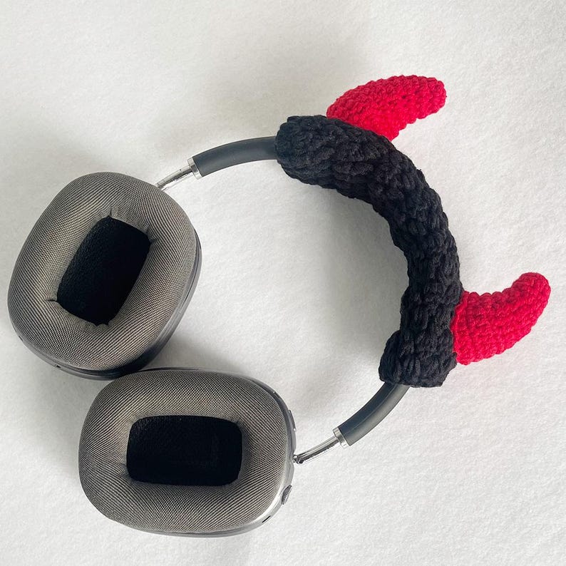 K&ouml;nnte beinhalten: Graue Kopfh&ouml;rer mit einem geh&auml;kelten Teufelshorn-Stirnband in Schwarz und Rot. Das Stirnband ist schwarz mit zwei roten H&ouml;rnern. Die Kopfh&ouml;rer haben graue Ohrmuscheln und ein silbernes Band.