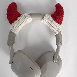 May include: A pair of gray headphones with red and white crocheted Santa hat ear covers. The ear covers are made of red and white yarn and are shaped like miniature Santa hats. The headphones are resting against a white background.