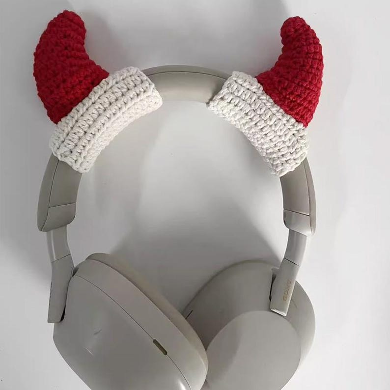 May include: A pair of gray headphones with red and white crocheted Santa hat ear covers. The ear covers are made of red and white yarn and are shaped like miniature Santa hats. The headphones are resting against a white background.