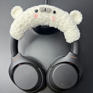 Puede incluir: Auriculares Sony negros con una funda blanca y esponjosa con forma de oso. La funda de oso tiene pequeños ojos negros, mejillas rosas y detalles grises. Los auriculares tienen el nombre de la marca "SONY" en color cobre.