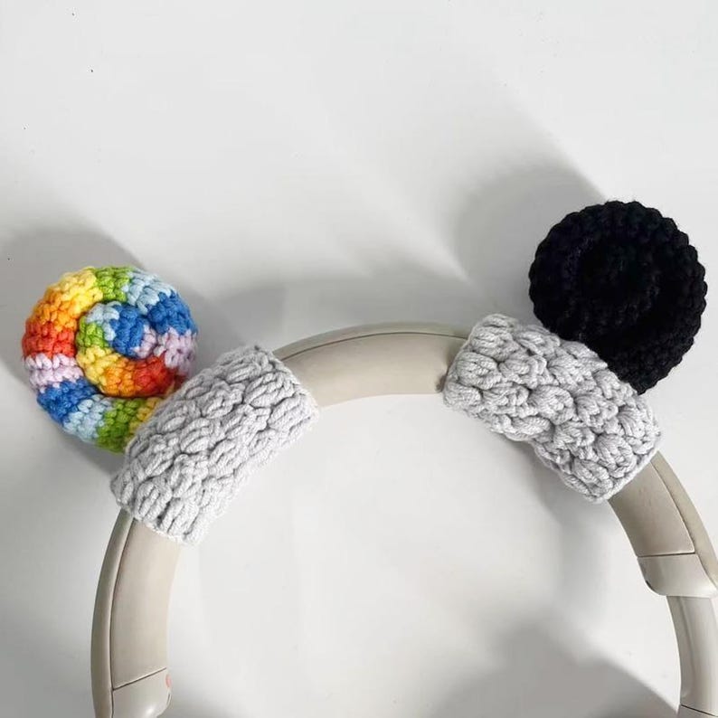 May include: Headphones with gray ear covers and decorative crochet accents. One accent is a rainbow spiral, and the other is a black circle. The headphones are set against a white background.