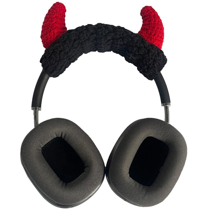 K&ouml;nnte beinhalten: Graue Over-Ear-Kopfh&ouml;rer mit schwarzem B&uuml;gel und Ohrmuscheln. Das Kopfband ist mit einer schwarzen, strukturierten Abdeckung und zwei roten, geh&auml;kelten Teufelsh&ouml;rnern verziert, ein einzigartiges Accessoire.
