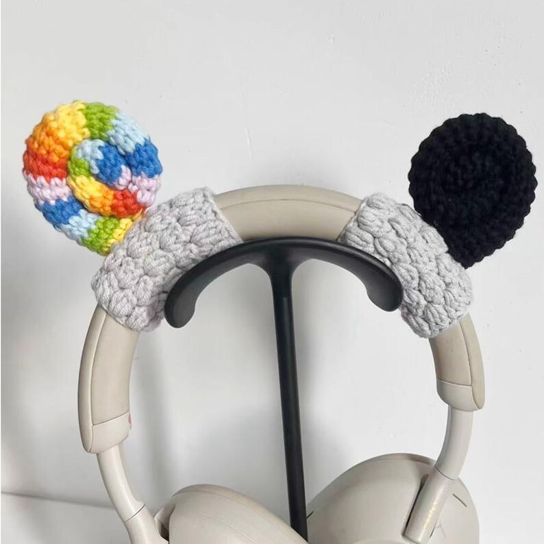 May include: Headphones with crocheted ear covers. One cover is a swirl of rainbow colors, the other is black. The headband is covered in gray crochet. The headphones are on a black stand.