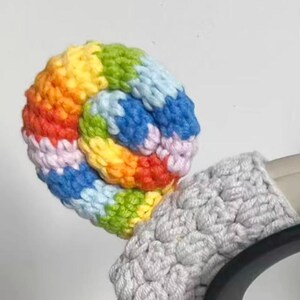 May include: A handmade, crocheted rainbow-colored ear warmer with a spiral design. The ear warmer is made of soft yarn in a variety of colors, including red, orange, yellow, green, blue, and purple. A gray crocheted band is attached to the ear warmer.