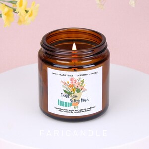 Puede incluir: Una vela en tarro de cristal marrón con una mecha encendida. La etiqueta dice "Thank You So Very Much" con una ilustración de ramo floral. La vela tiene aroma a Sea Salt Sage y un tiempo de combustión de 16 horas. La marca "FARICANDLE" está en la parte inferior.