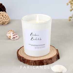Puede incluir: Una vela blanca encendida en un tarro de vidrio esmerilado con la inscripción "Barbara Bride to be". La vela está sobre un posavasos de madera. El texto "FARICANDLE" está en la parte inferior.