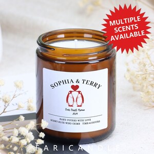 Op de afbeelding: Een bruine glazen kaarsenpot met een wit etiket met de namen "SOPHIA & TERRY", twee pinguïn illustraties en de tekst "Our First Home 2024". De kaars heeft een witte was en een brandtijd van 42 uur.