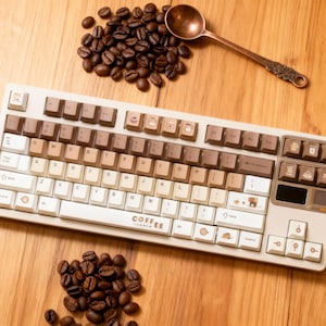 May include: A beige and brown keyboard with coffee-themed keycaps, coffee beans, and a decorative spoon on a wooden surface. The keyboard has the word "COFFEE" printed on it.