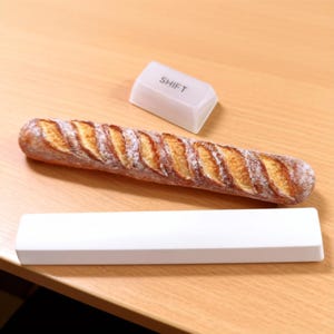 Könnte beinhalten: Ein knuspriges Baguette, eine weiße Tastaturtaste mit der Aufschrift "SHIFT" und ein weißes rechteckiges Objekt liegen auf einer Holzoberfläche. Das Brot ist goldbraun mit einer strukturierten Oberfläche.