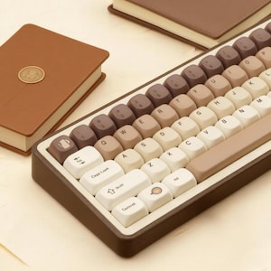 May include: A brown and cream-colored mechanical keyboard with a matching brown leather-bound notebook. The keyboard has a retro aesthetic with a variety of brown and cream keycaps. The notebook is closed and has a gold seal.