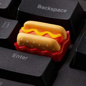 Può includere: Un copritasto a forma di hot dog, con panino giallo, hot dog rosso e senape gialla, è appoggiato su una tastiera nera. Il copritasto è in un vassoio rosso, aggiungendo un tocco giocoso alla tastiera.