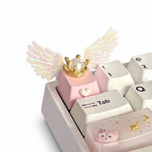 May include: A custom keyboard with a pink keycap featuring a white heart and a gold crown with a pearl. Iridescent wings are attached to the keycap. Other keys are white and clear with decorative elements.