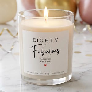 May include: A clear glass jar candle with a white label featuring the black text EIGHTY & Fabulous, AMAZING SINCE 1946, and a small red heart. The candle contains off-white wax, has a lit wick, and the label states soy wax candle 11 oz 312 g.