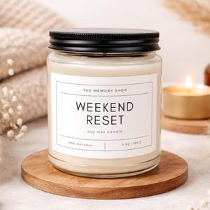 May include: A glass jar candle with a black lid and a white label that reads "WEEKEND RESET" in black text. The candle is on a wooden coaster. A lit tea light and a small brown bottle are in the background.