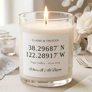 May include: A lit candle in a clear glass jar with a white label featuring the text CLAIRE & TRISTAN, 38.29687 N, 122.28917 W, Napa Valley - 07.20.2024, and Where It All Began. The candle wax is off-white and the flame is bright yellow.