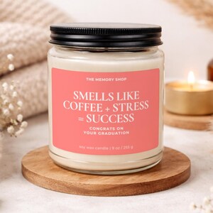 Peut inclure: Une bougie en pot de verre avec un couvercle noir et une étiquette rose qui dit "Smells Like Coffee + Stress = Success". La bougie est sur un dessous de verre en bois. La bougie est de 255 g.