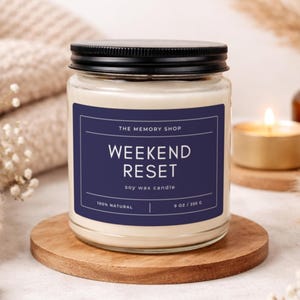 Könnte beinhalten: Ein Glas-Kerzenglas mit schwarzem Deckel und einem marineblauen Etikett. Auf dem Etikett steht "WEEKEND RESET" in weißer Schrift. Die Kerze steht auf einem Holzuntersetzer. Eine brennende Kerze und eine braune Flasche sind im Hintergrund.