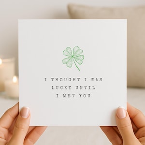 Puede incluir: Una tarjeta de felicitación blanca con una ilustración de trébol de cuatro hojas verde y el texto "I THOUGHT I WAS LUCKY UNTIL I MET YOU". La tarjeta está sostenida por manos, con velas encendidas en el fondo.