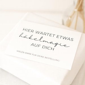 Könnte beinhalten: Ein weißes Paket mit einer weißen Karte oben drauf. Die Karte hat schwarzen Text mit der Aufschrift "HIER WARTET ETWAS häkelmagie AUF DICH VIELEN DANK FÜR DEINE BESTELLUNG!" Im Hintergrund eine Glasvase mit getrockneten Pflanzen.