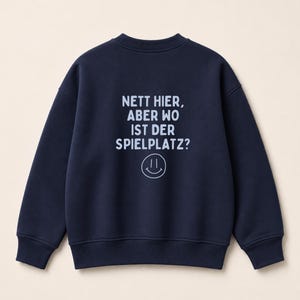 Könnte beinhalten: Ein dunkelblaues Langarm-Sweatshirt mit dem aufgedruckten Text 'NETT HIER, ABER WO IST DER SPIELPLATZ?' in hellblauer Schrift auf der Rückseite, ergänzt durch ein einfaches, linienförmiges Smiley-Gesicht unterhalb des Textes.