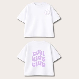 Könnte beinhalten: Zwei weiße T-Shirts. Die Vorderseite des oberen Shirts hat ein kleines, hellviolettes Smiley-Gesicht. Auf der Rückseite des unteren Shirts stehen die Worte "COOL KIDS CLUB" in hellvioletter, wellenförmiger Schrift. Die Shirts sind kurzärmelig.