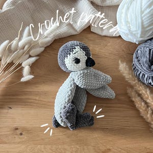 Op de afbeelding: Een handgemaakte gehaakte pinguïn in grijstinten, wit en zwart. De pinguïn heeft een afgerond lichaam, vleugels en poten. De tekst "Crochet pattern" is in cursief geschreven. Een bol witte wol en een bol grijze wol staan op de achtergrond.
