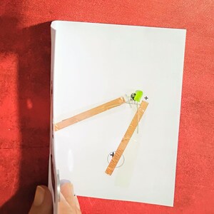 May include: A white sheet of paper with a simple circuit diagram. Copper tape forms the circuit, connecting a green LED to a battery symbol. A plus sign is next to the LED, and a minus sign is near the battery symbol. The background is red.