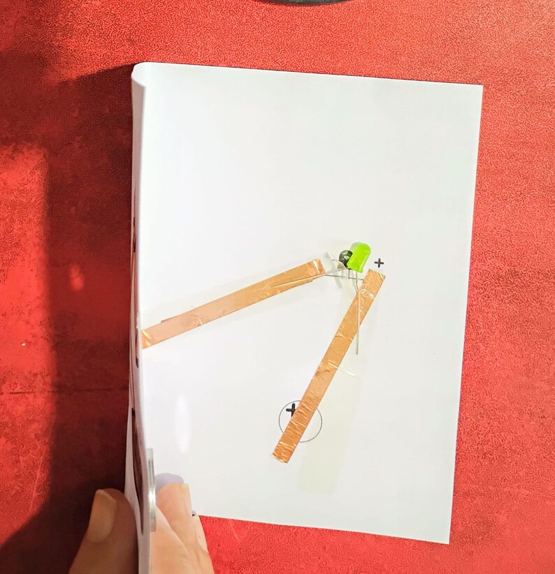 May include: A white sheet of paper with a simple circuit diagram. Copper tape forms the circuit, connecting a green LED to a battery symbol. A plus sign is next to the LED, and a minus sign is near the battery symbol. The background is red.