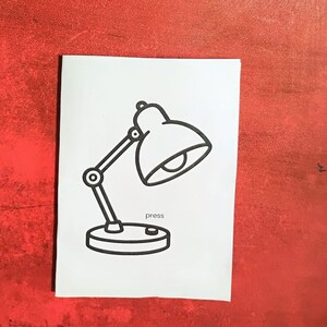 May include: A black and white line drawing of a desk lamp on a white background. The lamp has a rounded shade, an adjustable arm, and a circular base. The word "press" is written below the base.