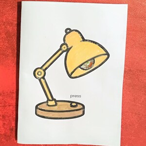 May include: A hand-drawn illustration of a yellow desk lamp with a brown base and arm. The word "press" is written below the lamp's base. The lamp's shade is angled downward, and the lightbulb is visible.