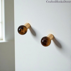 Puede incluir: Dos ganchos de pared decorativos con acabado de latón y una esfera redonda veteada marrón y dorada. Los ganchos están montados en una pared blanca. El texto "CraftedHooksDecor" está en la esquina superior derecha.