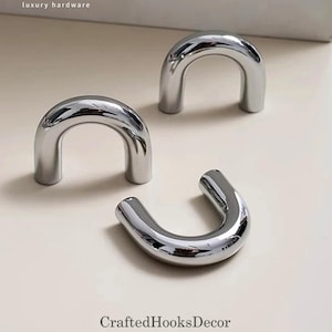 May include: Three shiny, silver-colored, U-shaped cabinet pulls. The pulls are arranged on a light-colored surface. The text "luxury hardware" is at the top of the image, and "CraftedHooksDecor" is at the bottom.