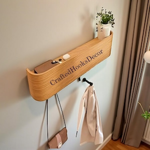 Peut inclure: Étagère murale en bois incurvée, avec une petite plante en pot, des clés et un portefeuille. Un sac à main et un manteau sont suspendus en dessous. Le texte "CraftedHooksDecor" est imprimé sur l'étagère.