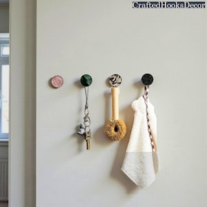 May include: A collection of decorative wall hooks in various colors and patterns. One hook is pink, another is green, a third has a speckled design, and the last is black. Items such as keys, a brush, and a towel are hanging from the hooks. The text "CraftedHooksDecor" is at the top.
