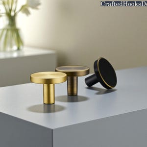 Może przedstawiać: Trzy dekoracyjne gałki do szafek w kolorze złotym, brązowym i czarnym ze złotym wykończeniem. Gałki mają cylindryczną podstawę i płaski, okrągły wierzch. Gałki są ułożone na szarej powierzchni. Tekst "CraftedHooksDecor" znajduje się w prawym górnym rogu.