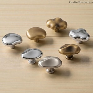 May include: A collection of mushroom-shaped cabinet knobs in various metallic finishes, including silver, gold, and bronze. The knobs have a smooth, rounded design and are arranged on a light wood surface. The text "CraftedHooksDecor" is visible in the top right corner.